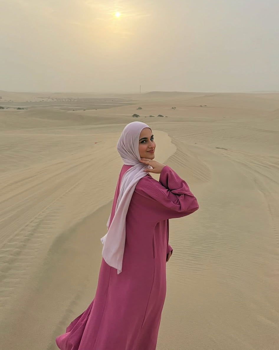 A woman wearing a pink abaya and a light pink hijab stands on a desert sand dune, looking back over her shoulder towards the camera under a hazy sunset sky.