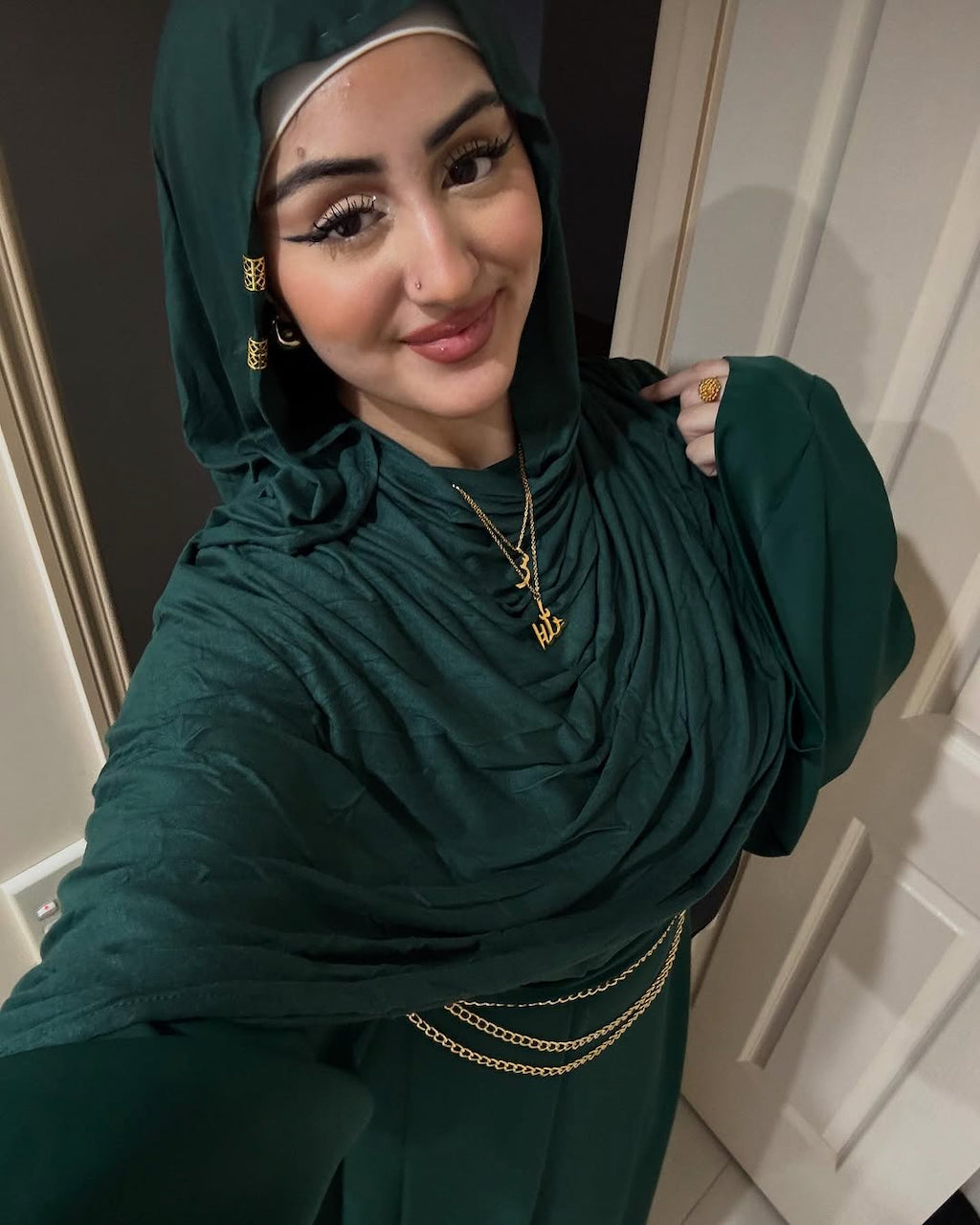 A woman wearing a dark green draped hijab and matching abaya with gold chain accents, taking a mirror selfie in an indoor setting.