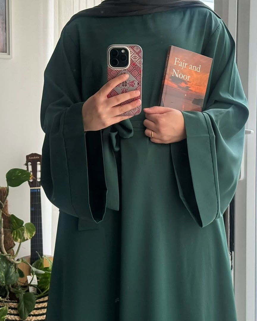 Person wearing a green abaya holding a phone and a book indoors.