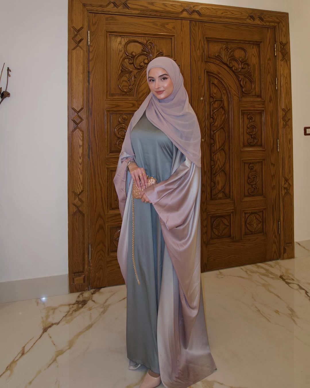 Woman in a long, light-colored dress with a matching hijab standing in front of a wooden door.
