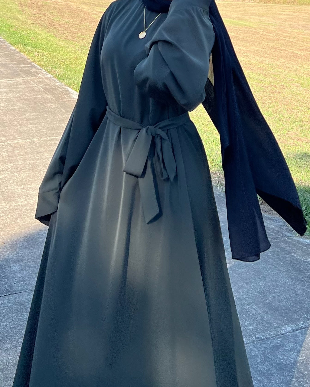 A woman wearing a dark navy blue abaya dress with a matching headscarf and a self-tie waist belt, standing outdoors on a paved path with grass in the background.