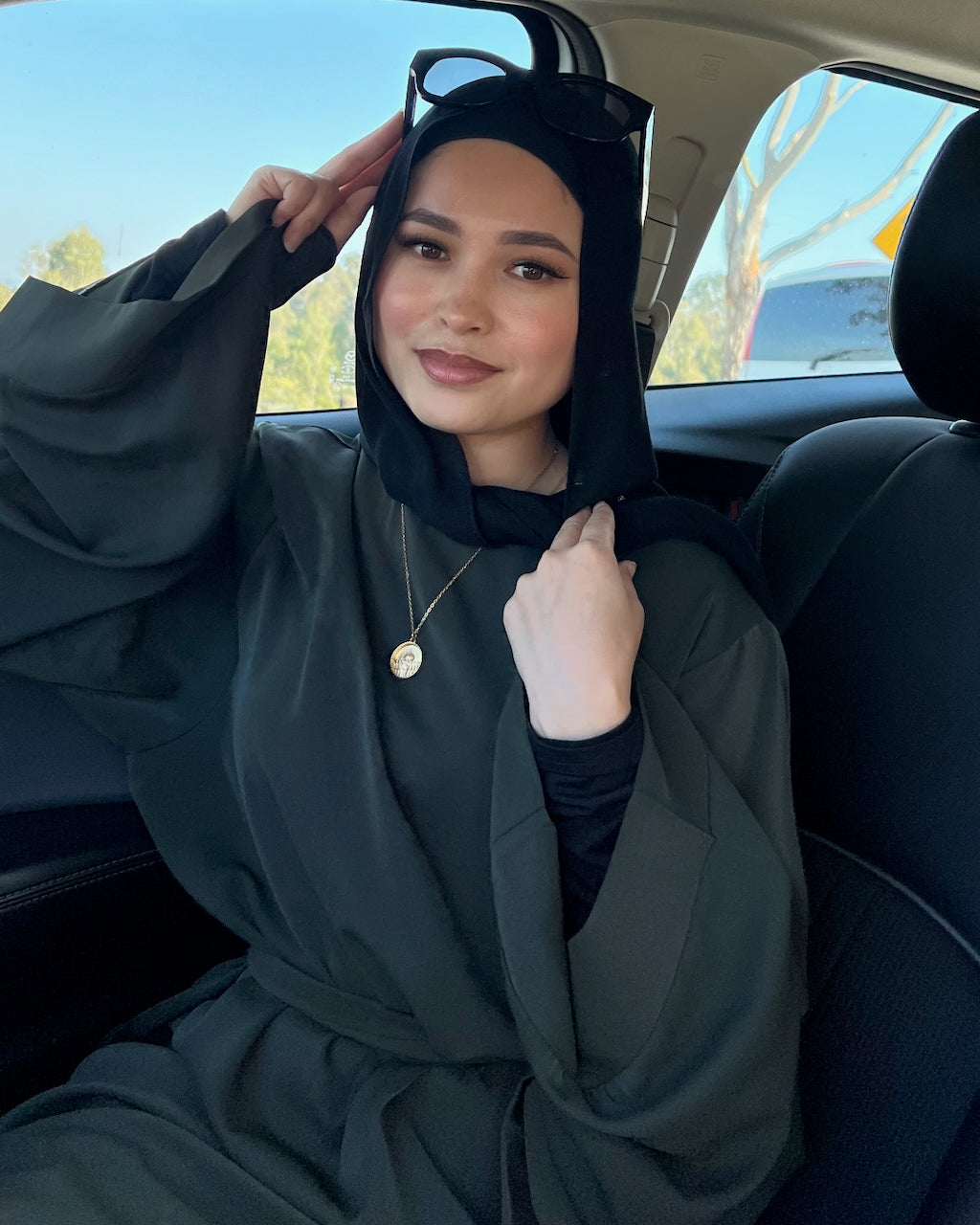 A woman wearing a dark olive green hijab and matching long-sleeve abaya sits in the back seat of a car, holding her hijab up with one hand.