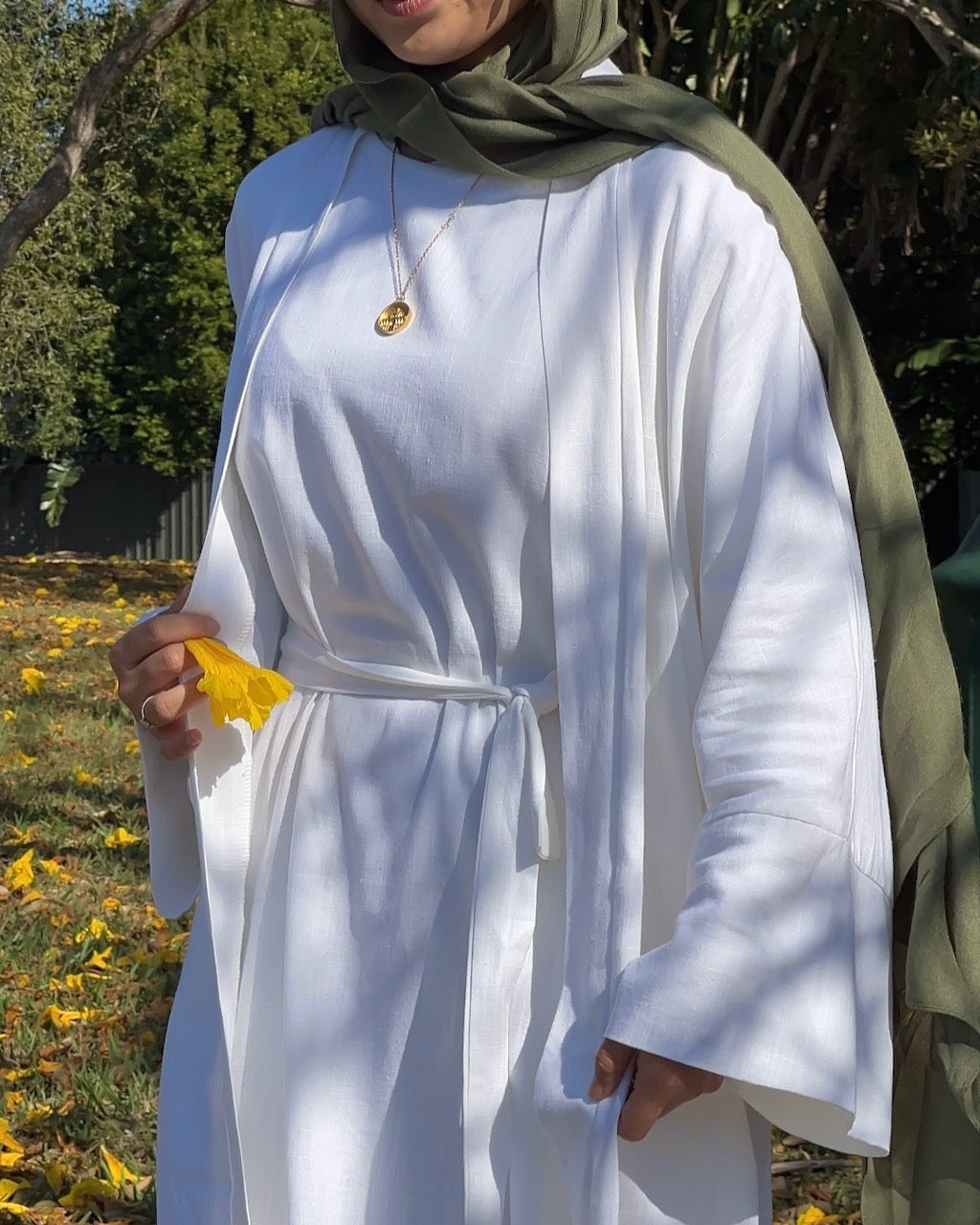 A woman outdoors wearing a white long-sleeve tunic with a tie waist and an olive green headscarf, holding a yellow leaf. The background shows trees and fallen leaves.