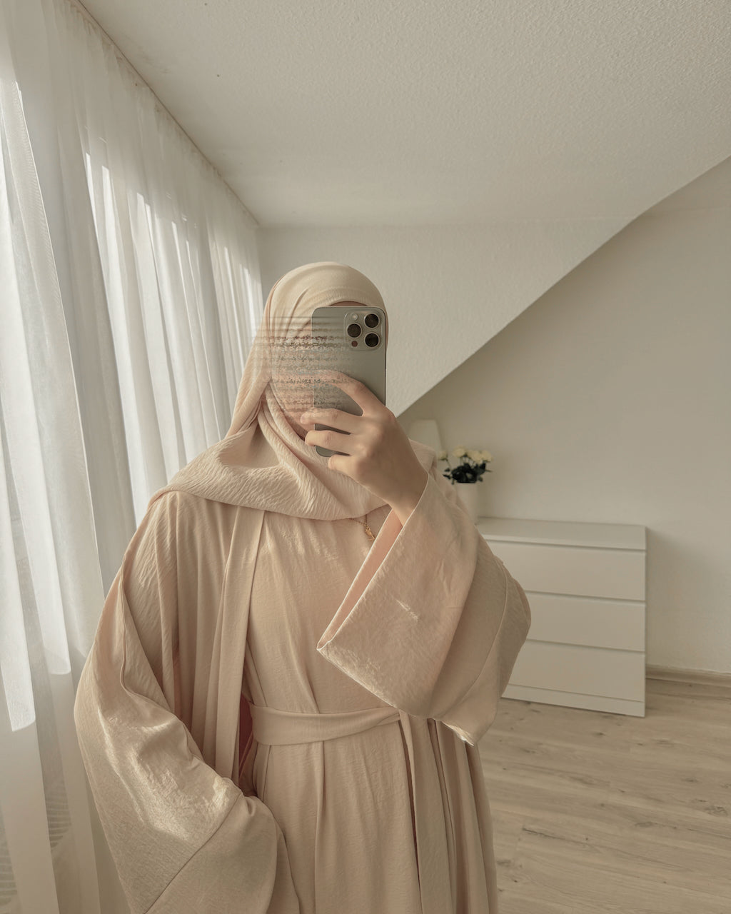 A person taking a mirror selfie in a minimalist room, wearing a light beige hooded robe with wide flowing sleeves and a matching belt.