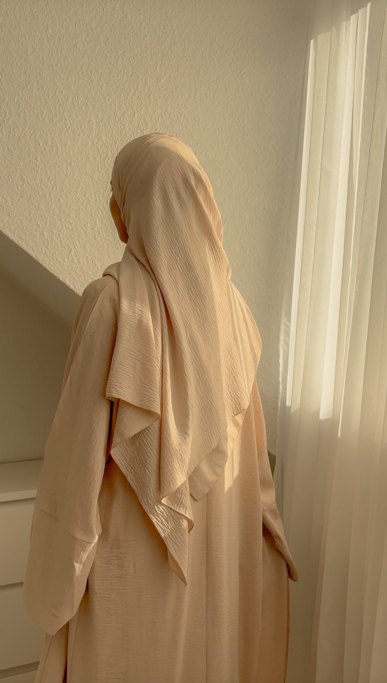 Person wearing a beige hijab and dress standing in a softly lit room.