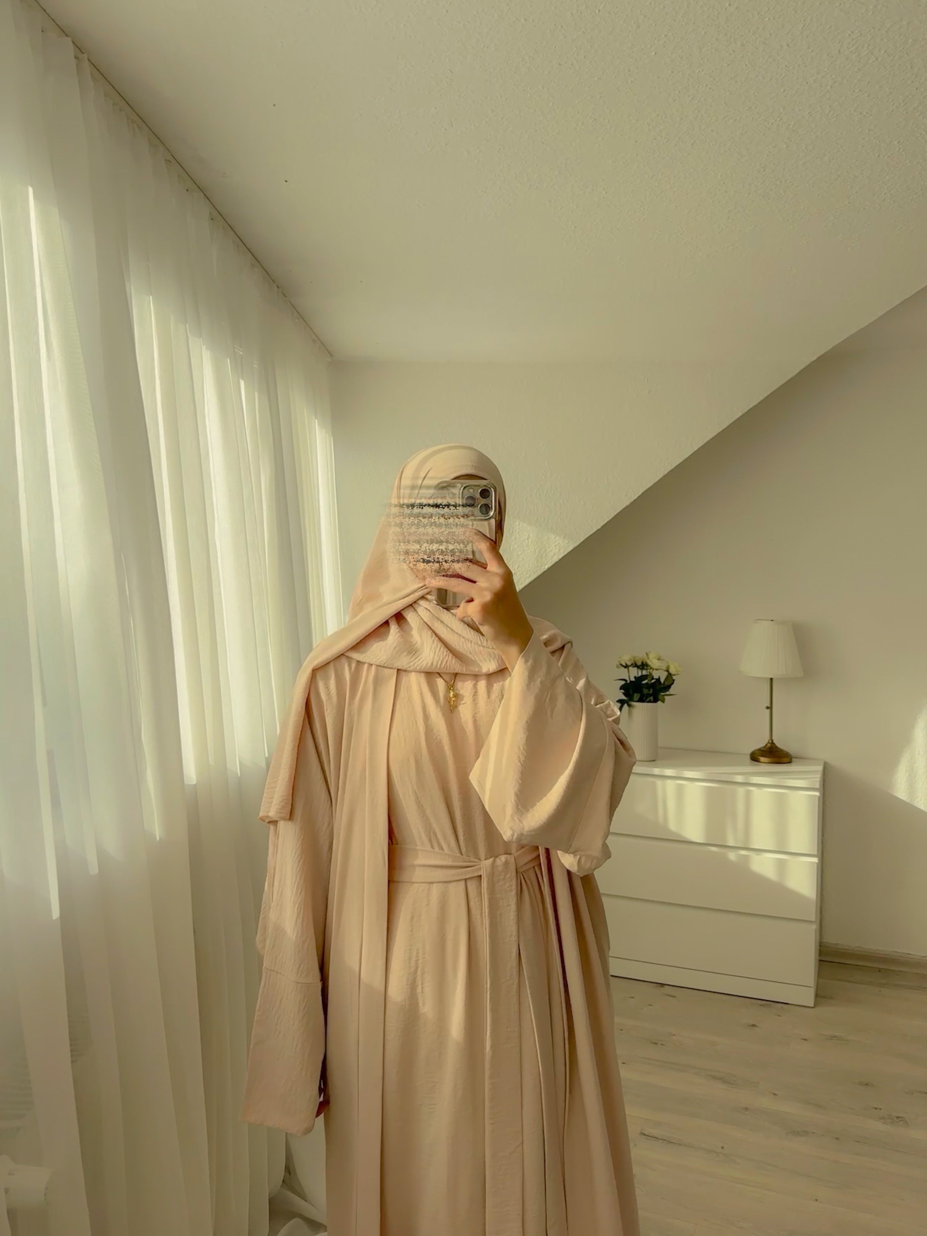 Person wearing a beige outfit in a room with white curtains and a dresser.
