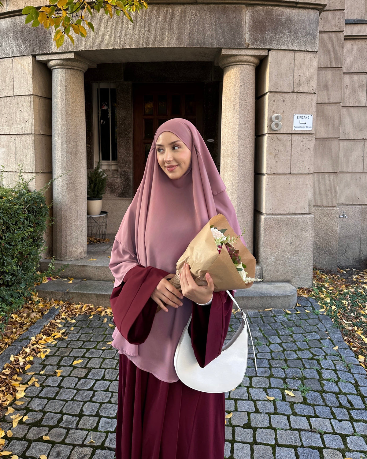 A woman wearing a dusty rose hijab and a deep burgundy maxi dress stands outdoors in front of a stone building entrance, holding a brown paper-wrapped bouquet and a white shoulder bag.