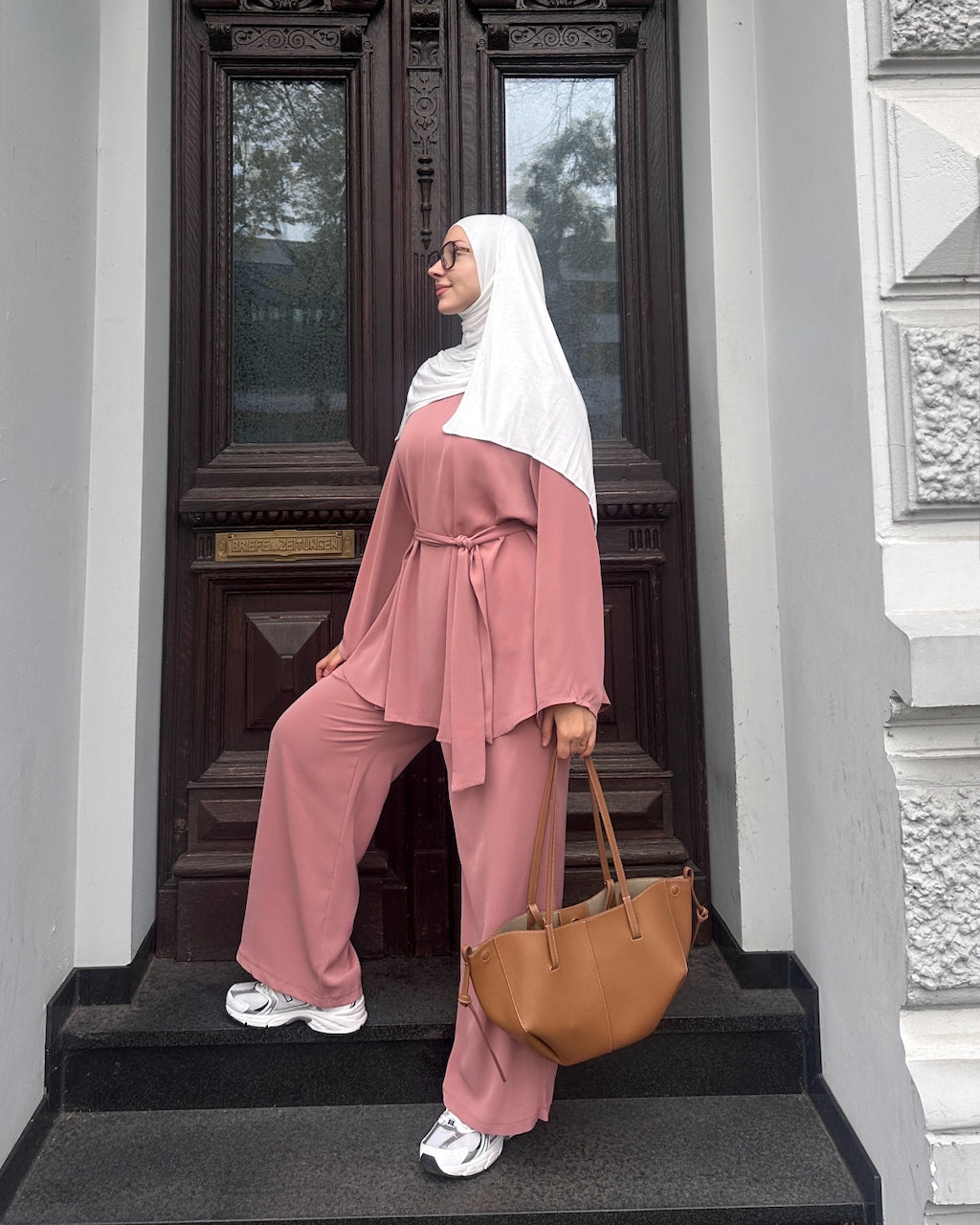 A woman wearing a dusty pink two-piece abaya set with a white hijab stands on black steps in front of a dark wooden door. She is holding a tan leather tote bag.