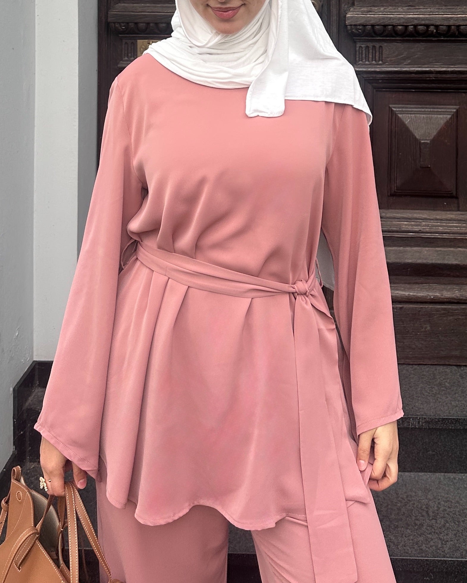 Person wearing a pink outfit with a white hijab standing indoors.
