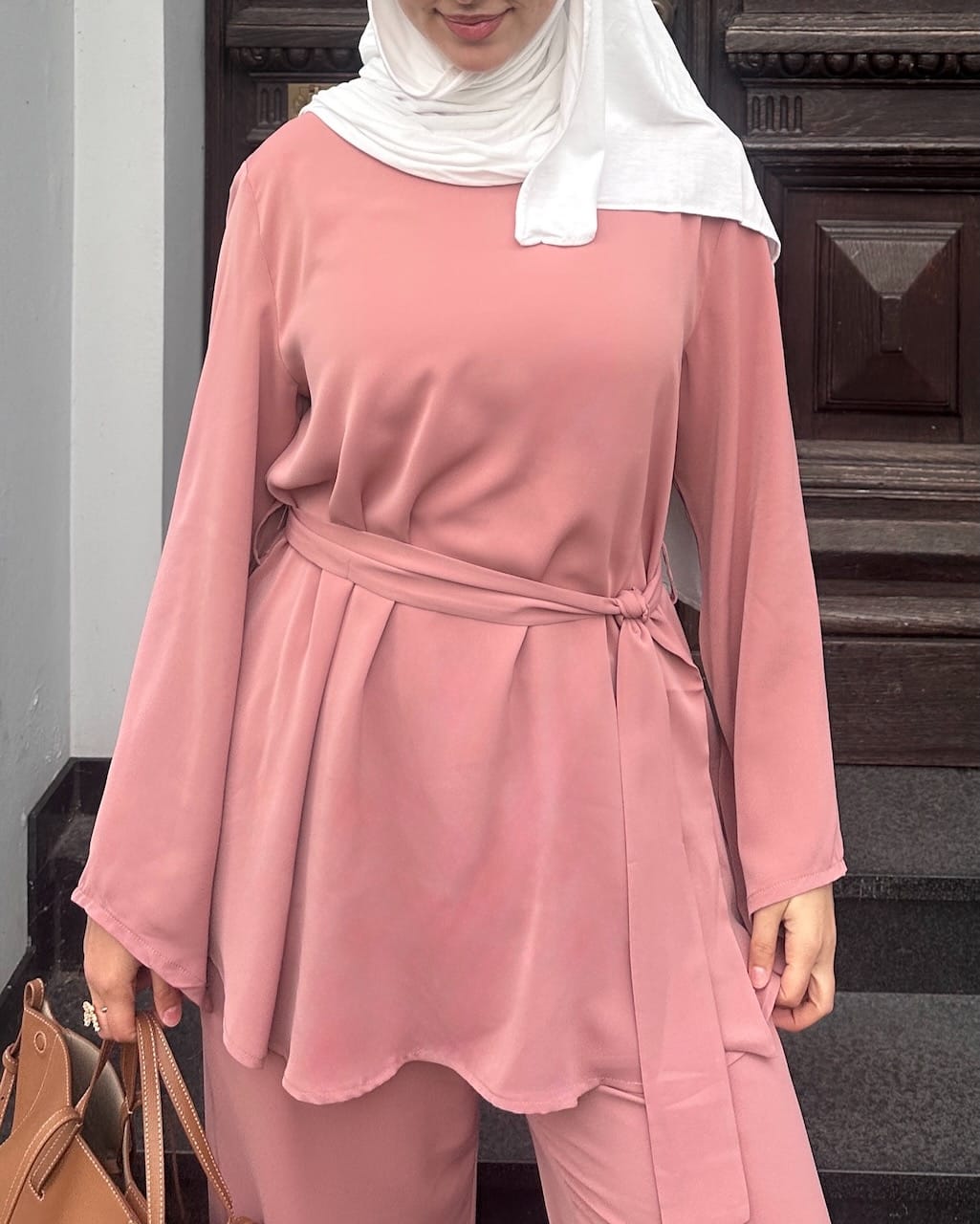 A woman wearing a dusty pink long-sleeve modest tunic with a matching tie belt and matching trousers, paired with a white hijab and holding a brown handbag.