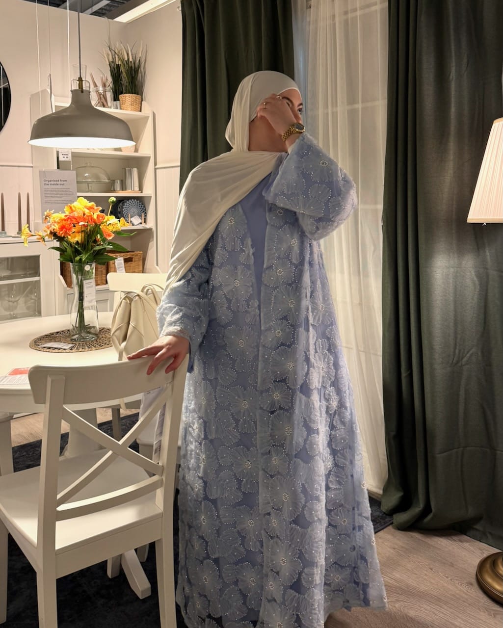 A woman wearing a light blue floral embroidered abaya and a white hijab stands indoors near a dining table and chairs, holding a phone to her face.