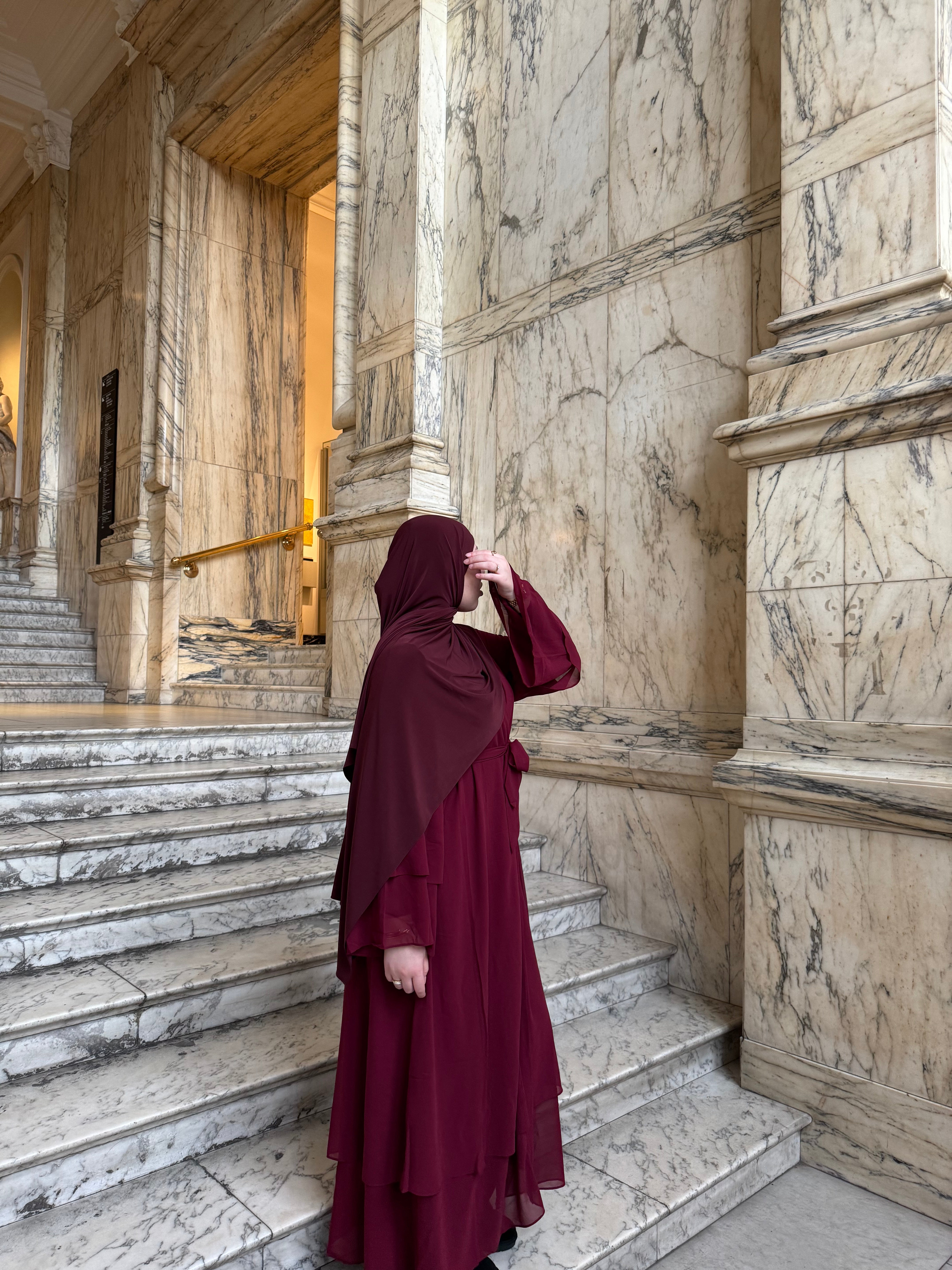 Woman in a burgundy hijab and dress standing on marble steps inside a grand building.
