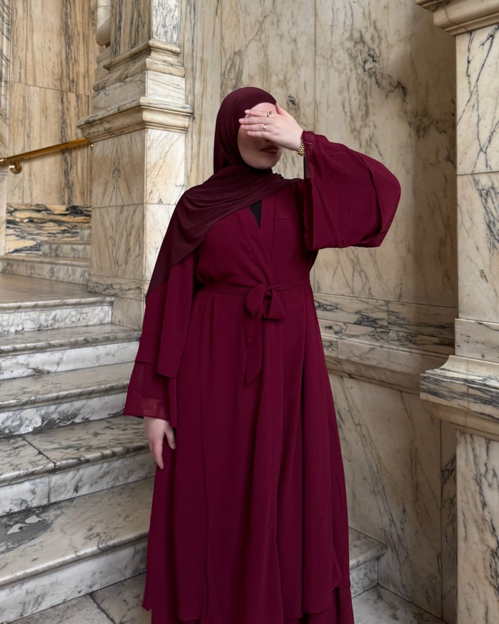 A woman wearing a long burgundy abaya dress with a matching hijab stands on marble stairs, covering part of her face with her hand.