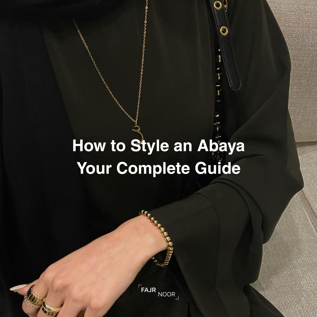 A close-up lifestyle shot of a person wearing a black abaya with a gold chain necklace and a gold beaded bracelet, with white text overlay reading 'How to Style an Abaya Your Complete Guide'.