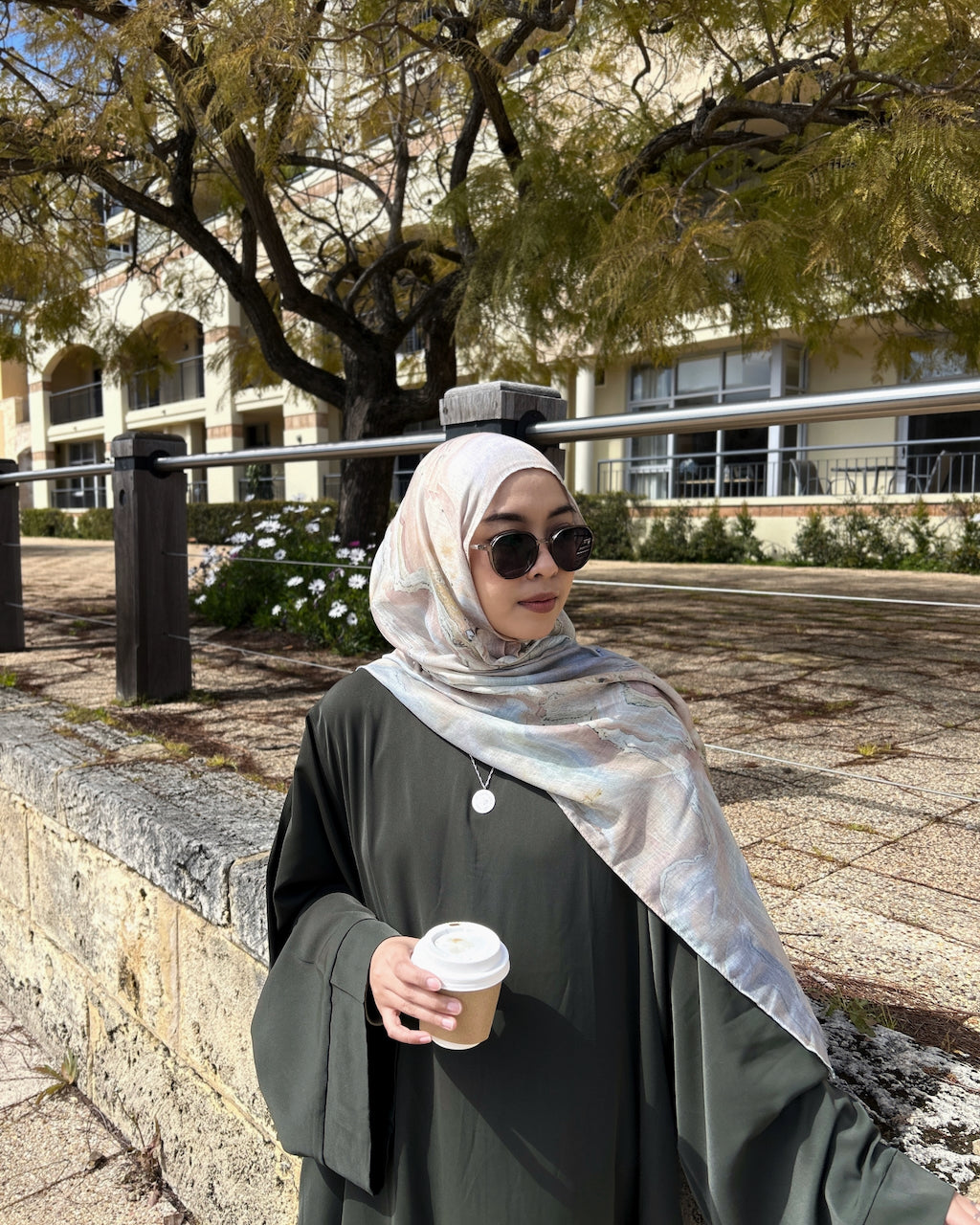 A woman wearing a dark olive green abaya and a light beige patterned hijab stands outdoors holding a coffee cup. She is wearing round black sunglasses, and the background features a stone wall, trees, and a building.