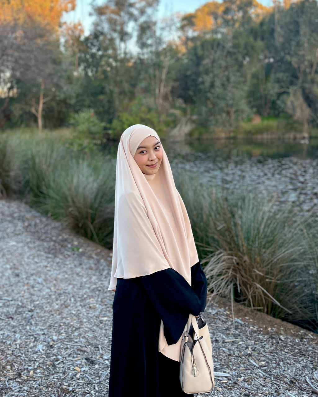 A woman wearing a light beige hijab and a black abaya stands on a gravel path outdoors near a pond with trees in the background, holding a cream-colored handbag.