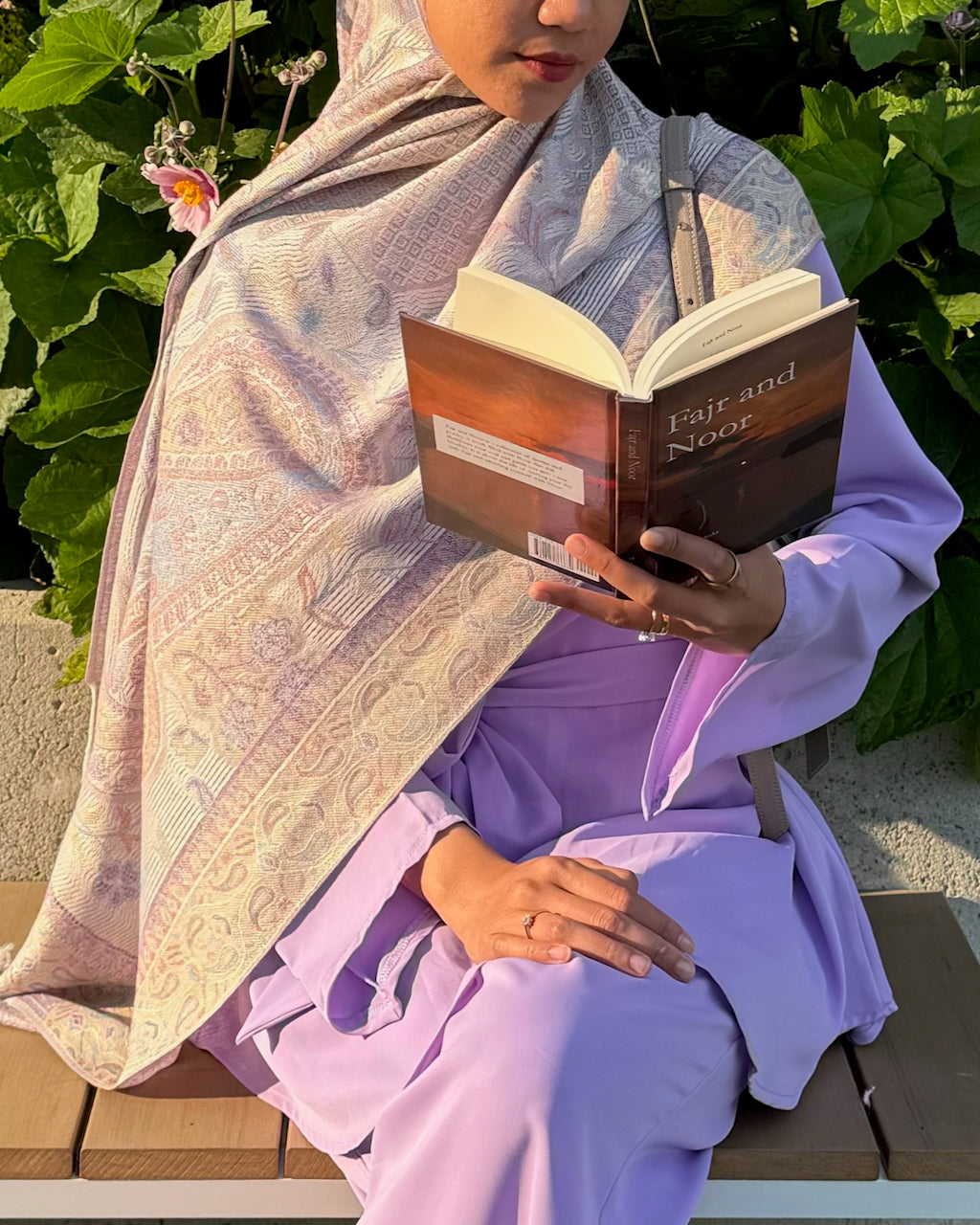 A woman in a lavender abaya and matching hijab sits on a wooden bench outdoors, holding an open book titled 'Fair and Narrow'. The background features lush green garden foliage.