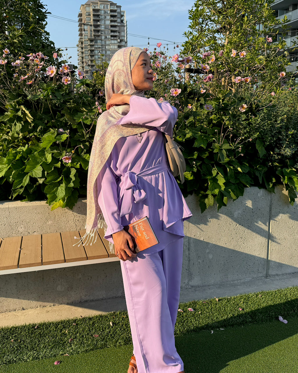 A woman wearing a lavender two-piece matching set with a tie waist and matching headscarf stands outdoors in front of green bushes and a bench.