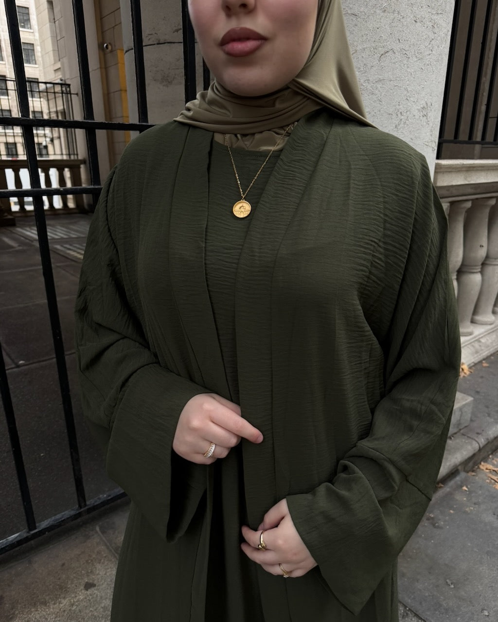 A woman wearing an olive green textured abaya with a matching hijab and a gold pendant necklace, standing outdoors in front of a stone wall and black metal gate.
