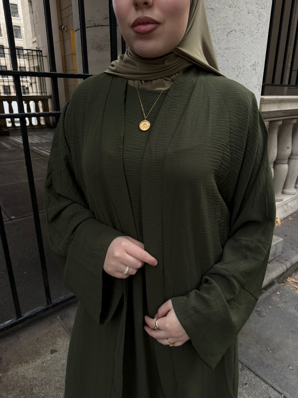 A woman wearing an olive green textured long-sleeve abaya with a matching hijab, standing outdoors in front of a stone wall and black metal gate.