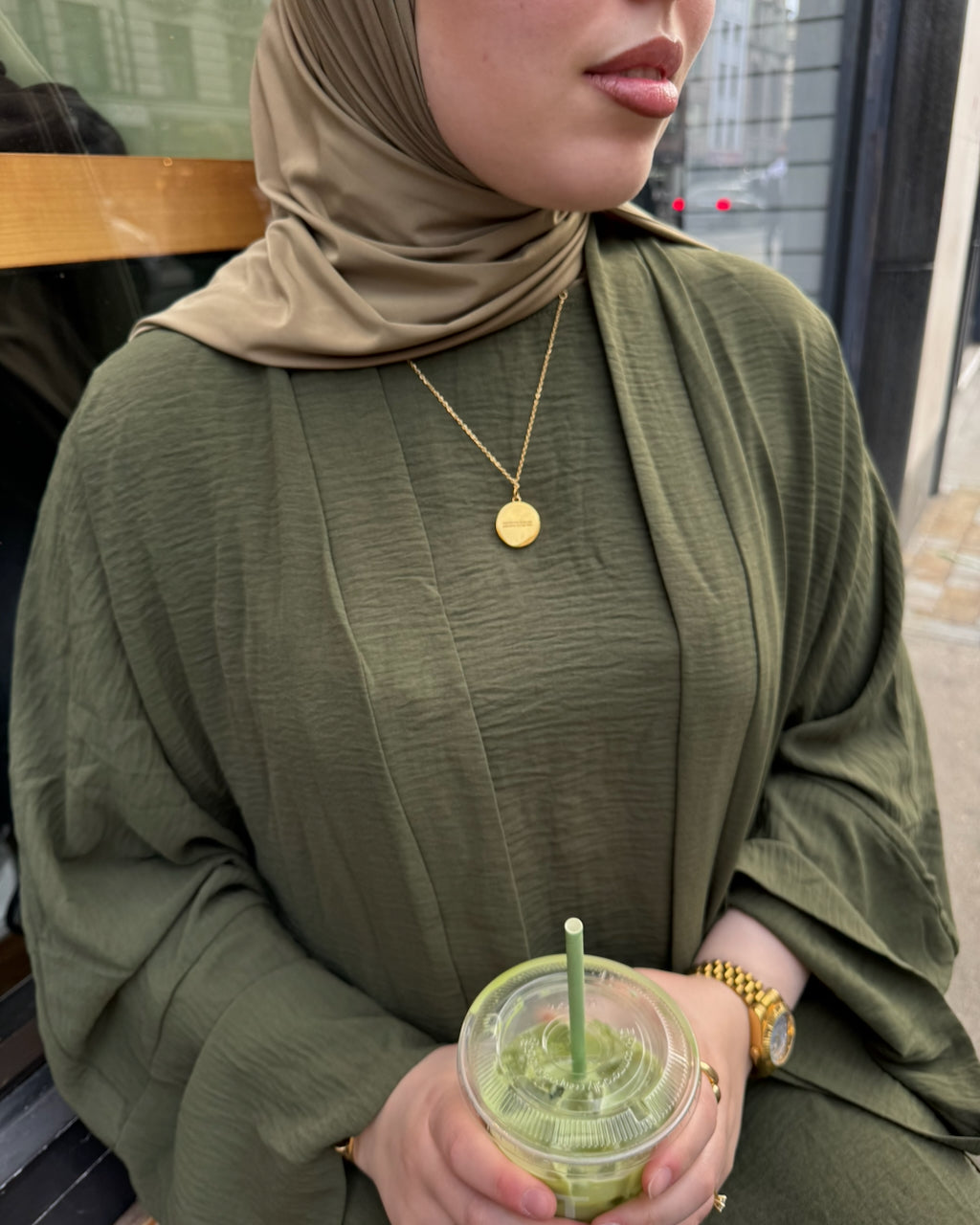 A woman wearing an olive green crinkled abaya and matching hijab, holding a green iced coffee. She is wearing a gold coin necklace and a gold watch.