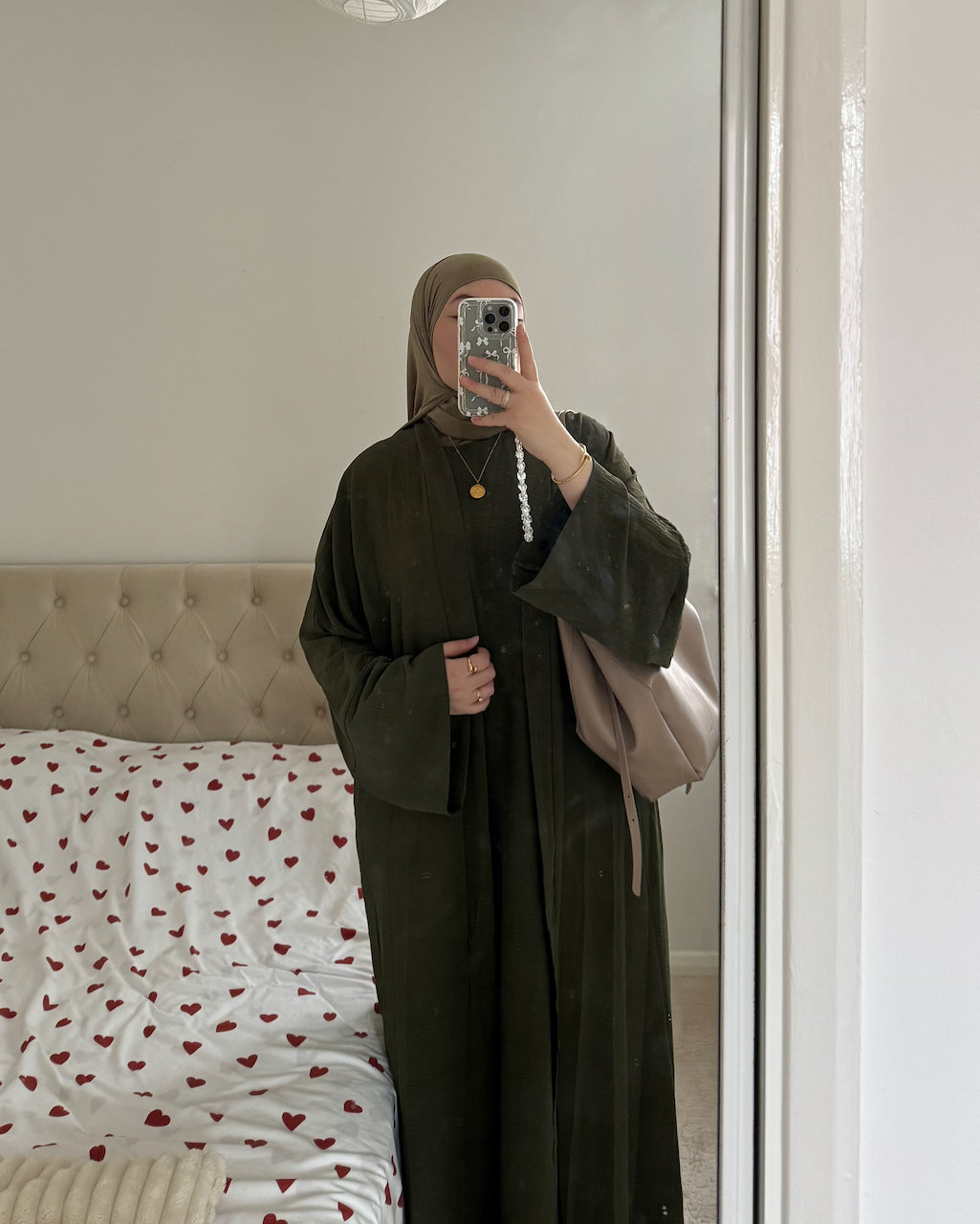 A woman takes a mirror selfie in a bedroom wearing a dark olive green abaya with wide sleeves and a matching headscarf. She is holding a phone in front of her face and carrying a light beige shoulder bag.