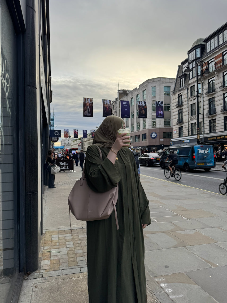 A woman wearing a dark olive green abaya and matching hijab stands on a london city sidewalk, holding a takeaway coffee cup and carrying a large dusty pink shoulder bag. The background shows a busy urban street scene with buildings, cars, and banners.