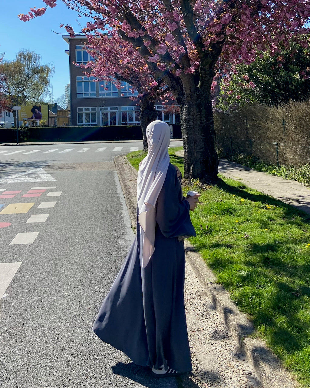A woman wearing a long dark gray abaya and a light beige hijab stands on a sunlit street beside a grassy verge and a blooming pink cherry tree, photographed from behind.