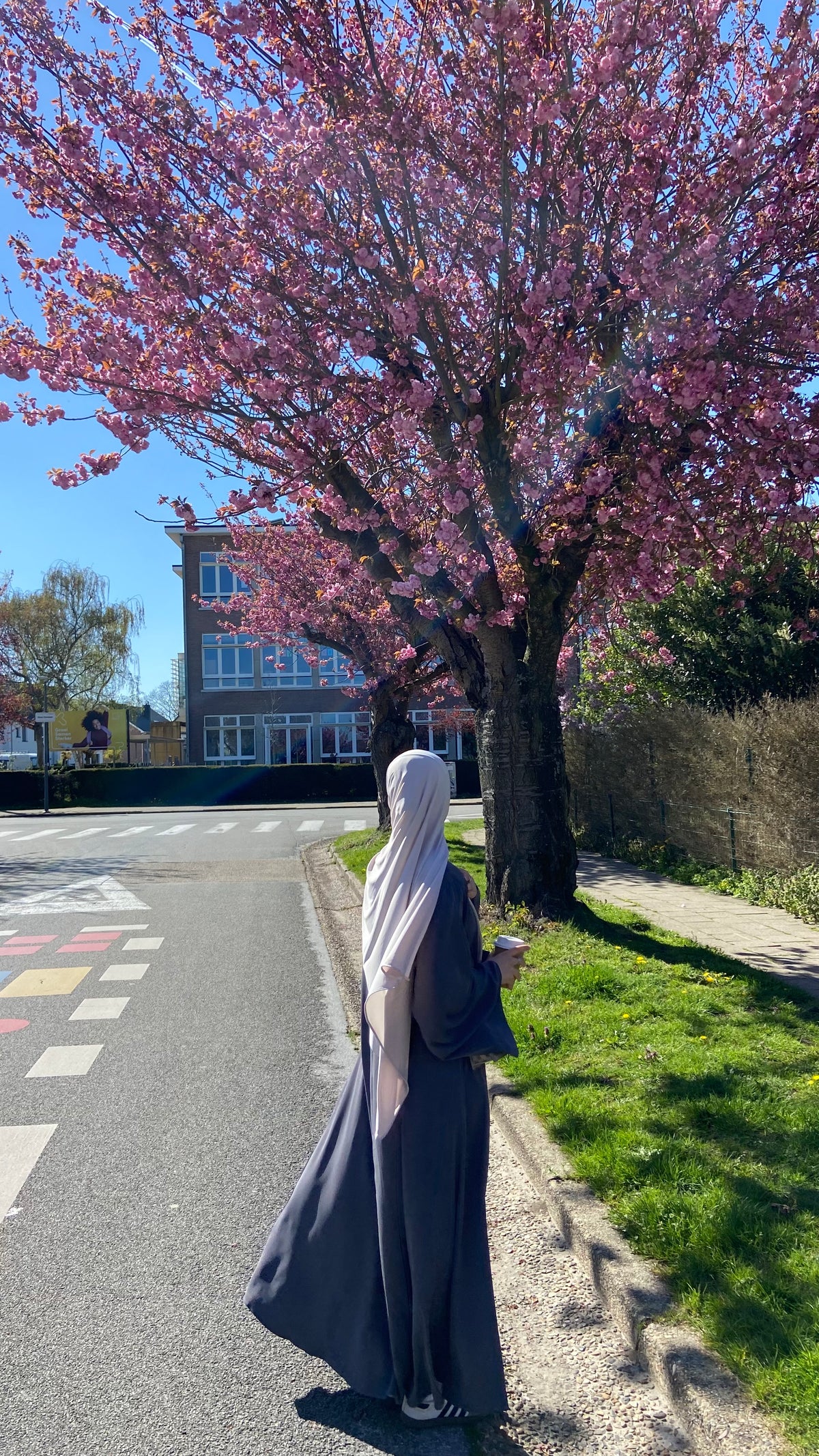 Person in a long coat and headscarf standing under a pink flowering tree on a sunny day.