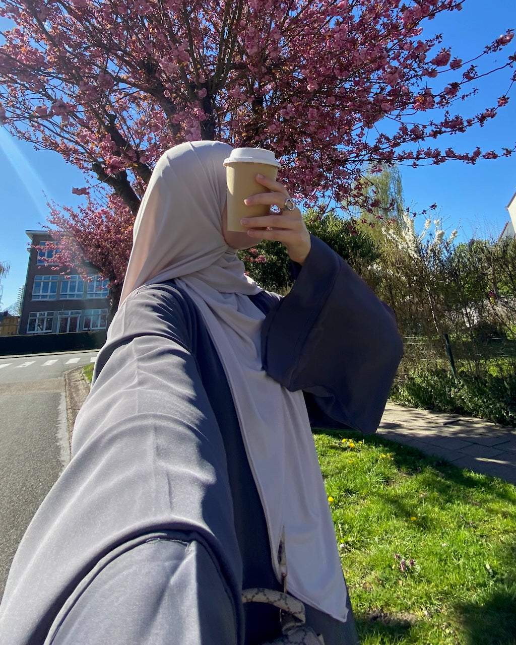 A person outdoors wearing a light beige hijab and a dark gray long-sleeve top, holding a takeaway coffee cup in front of their face. A pink flowering tree and a blue sky are visible in the background.