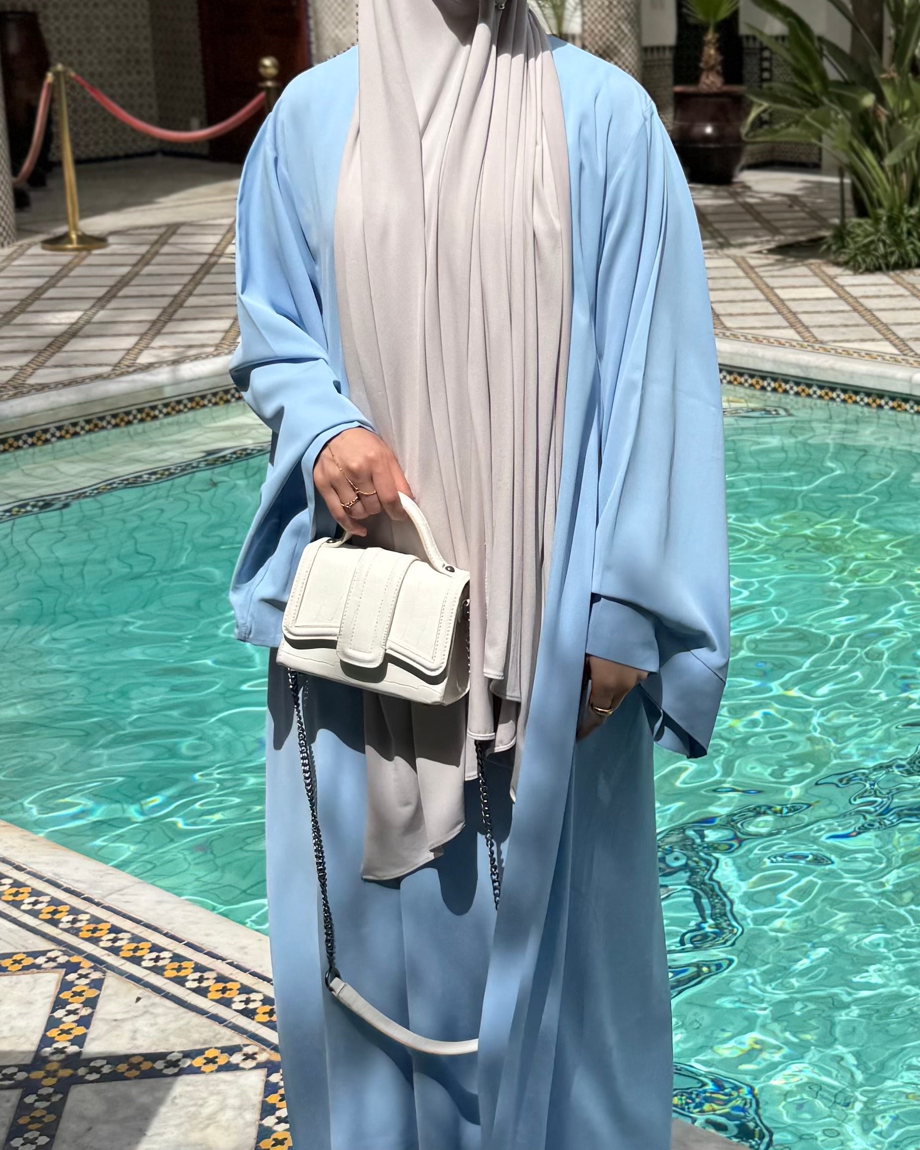 Person wearing a light blue outfit with a white hijab and holding a white handbag by a pool.