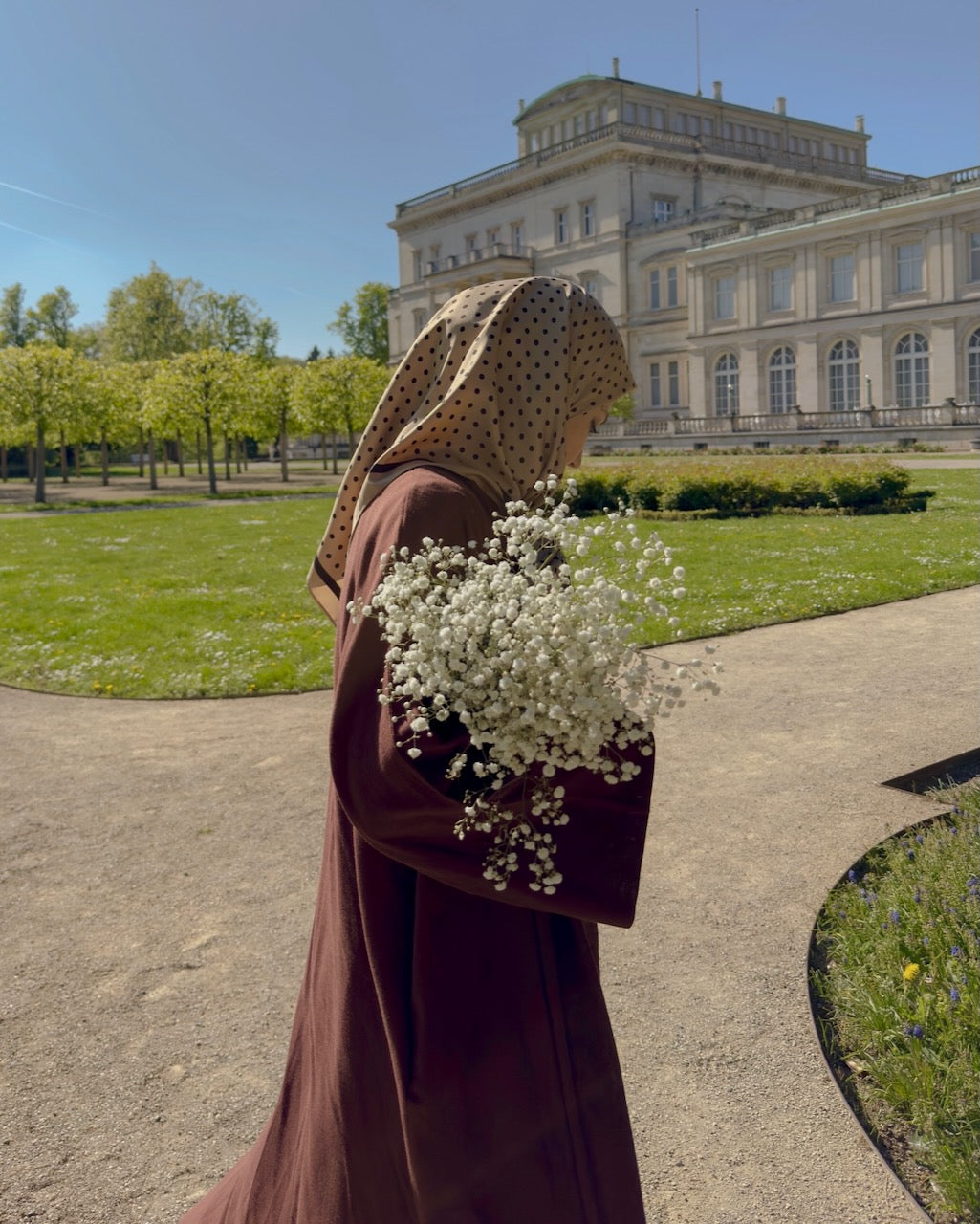A woman in a brown robe and patterned headscarf stands outdoors in a park holding a bouquet of white baby's breath, with a large classical building in the background.