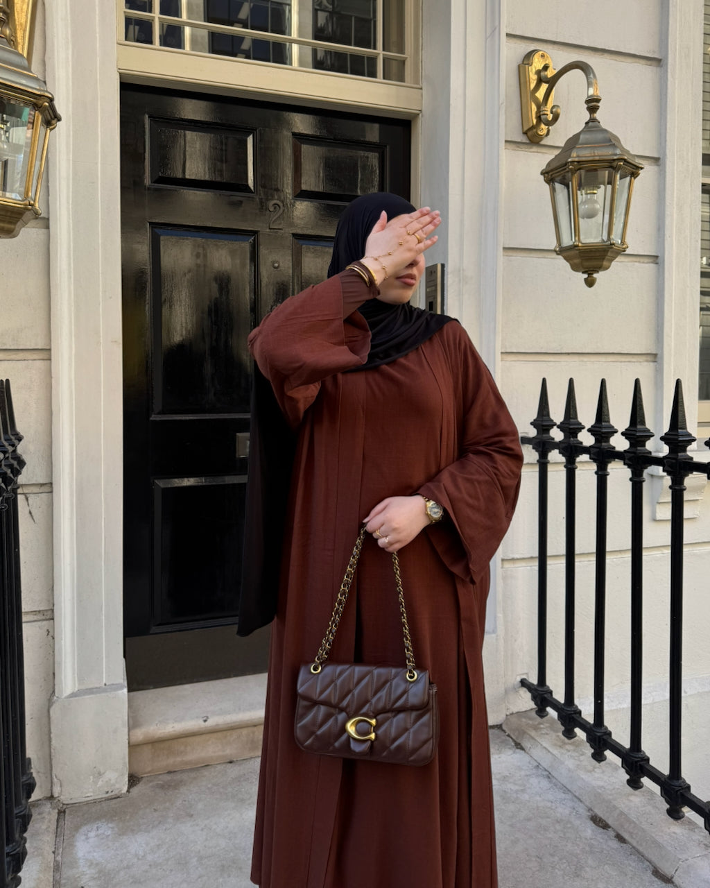 A woman wearing a long brown coffee linen abaya and black hijab stands in front of a black door and cream-colored building facade, holding a brown quilted handbag with a gold chain strap.