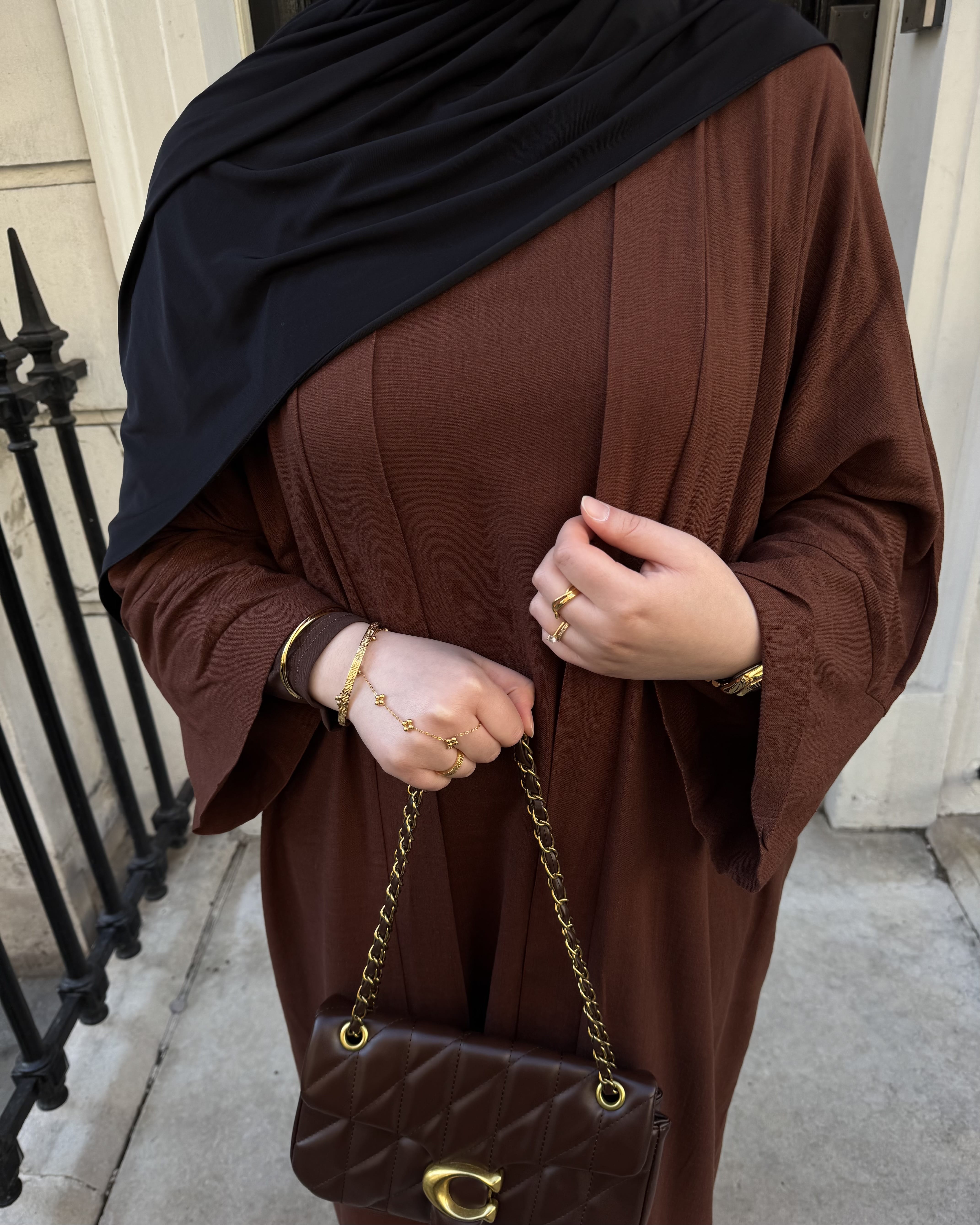 A person wearing a brown abaya and black hijab, holding a brown quilted shoulder bag with a gold chain strap and C-shaped clasp, standing outdoors near a black metal fence.