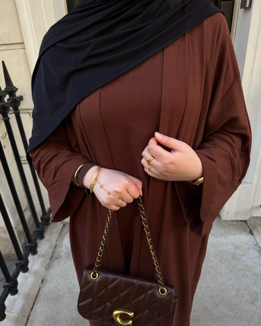 A person wearing a brown long-sleeve abaya or dress with a black hijab, holding a brown quilted shoulder bag with a gold chain strap in an outdoor urban setting.