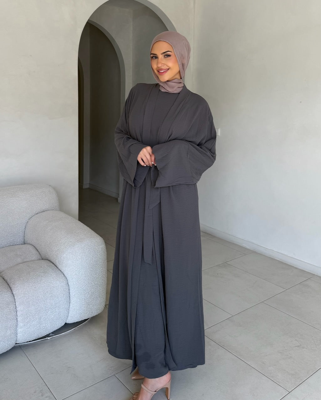A woman wearing a dark gray abaya and matching hijab stands in a minimalist room with light gray tiled floors and a curved white wall archway. The abaya has a loose, flowing silhouette with wide sleeves and a self-tie waist belt.