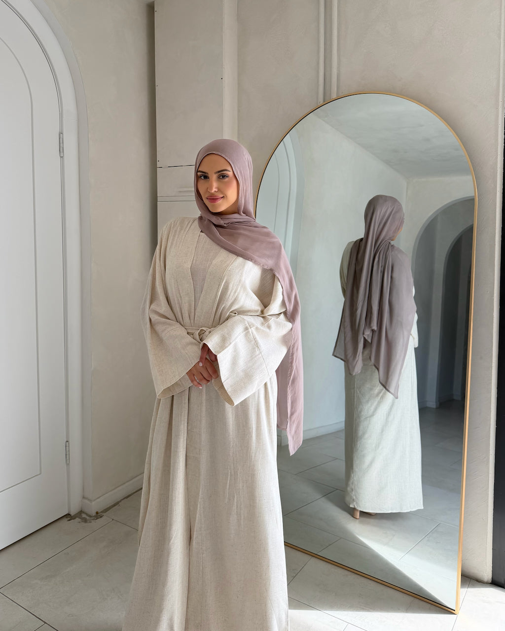 A woman wearing a sand linen abaya and a dusty mauve hijab stands in a minimalist room with a large arched mirror, showing both her front and side/back views in the reflection.