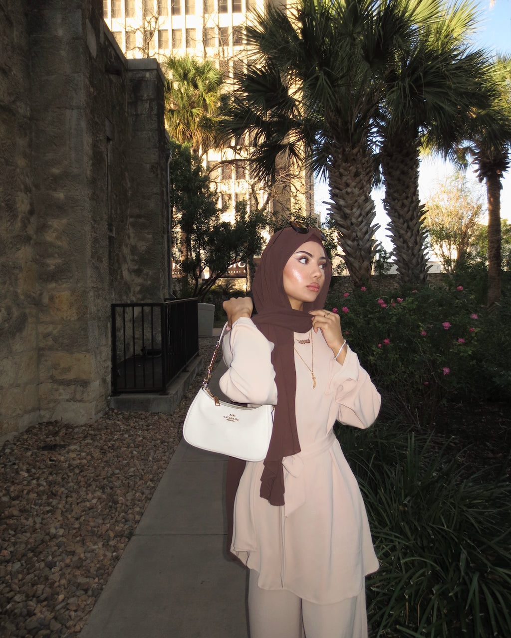 A woman wearing a light beige tunic and matching pants with a dark brown hijab, carrying a white handbag, standing outdoors on a sidewalk next to a stone wall and palm trees.
