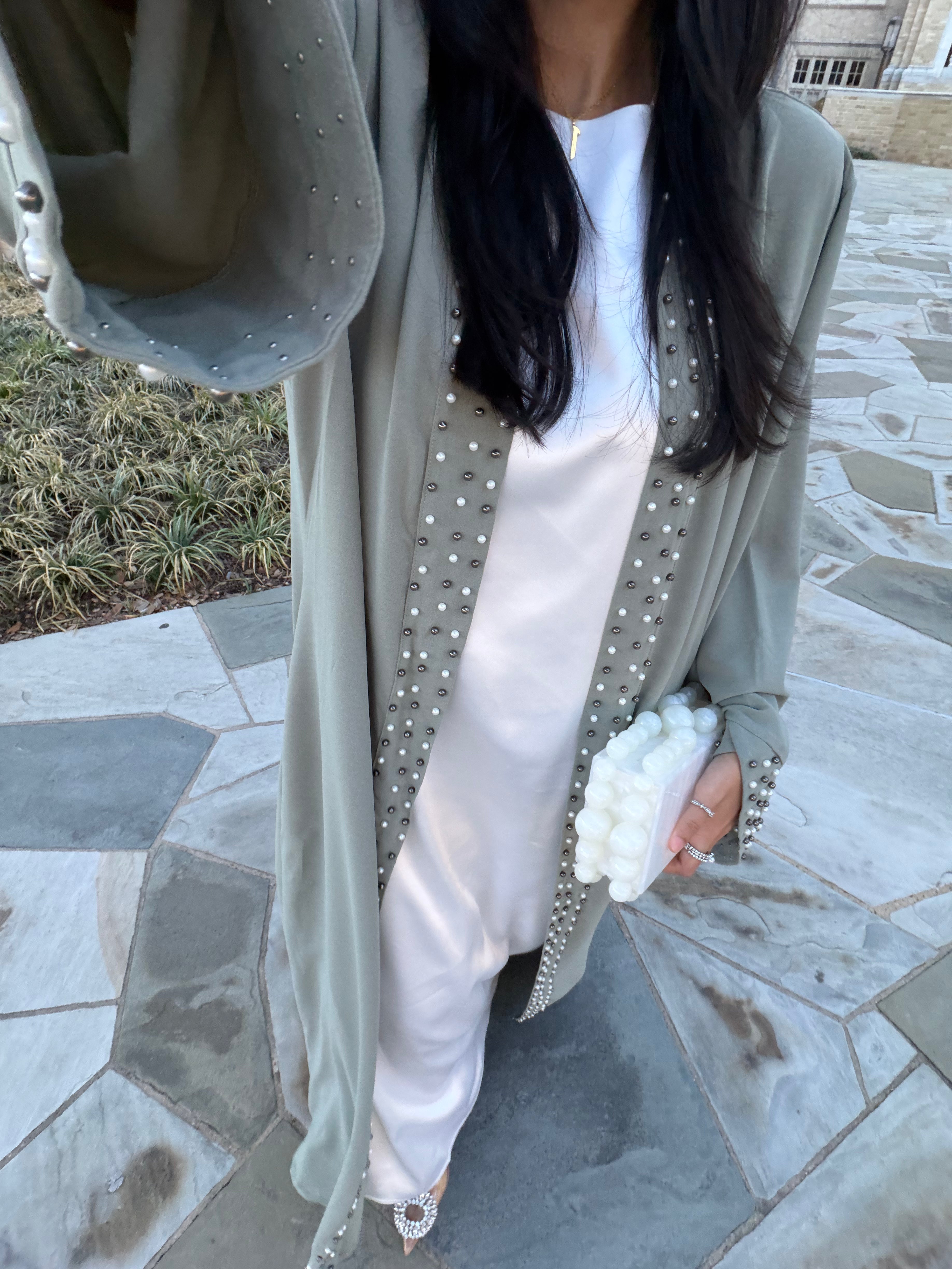 A woman wearing a long sage green open-front robe-style outer layer with silver beaded trim over a white satin dress, holding a white textured clutch on a stone patio.