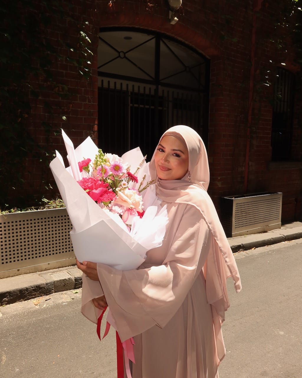 A woman wearing a light pink hijab and matching long-sleeve dress stands outdoors on a street holding a large bouquet of pink and white flowers wrapped in white paper, with a brick building in the background.