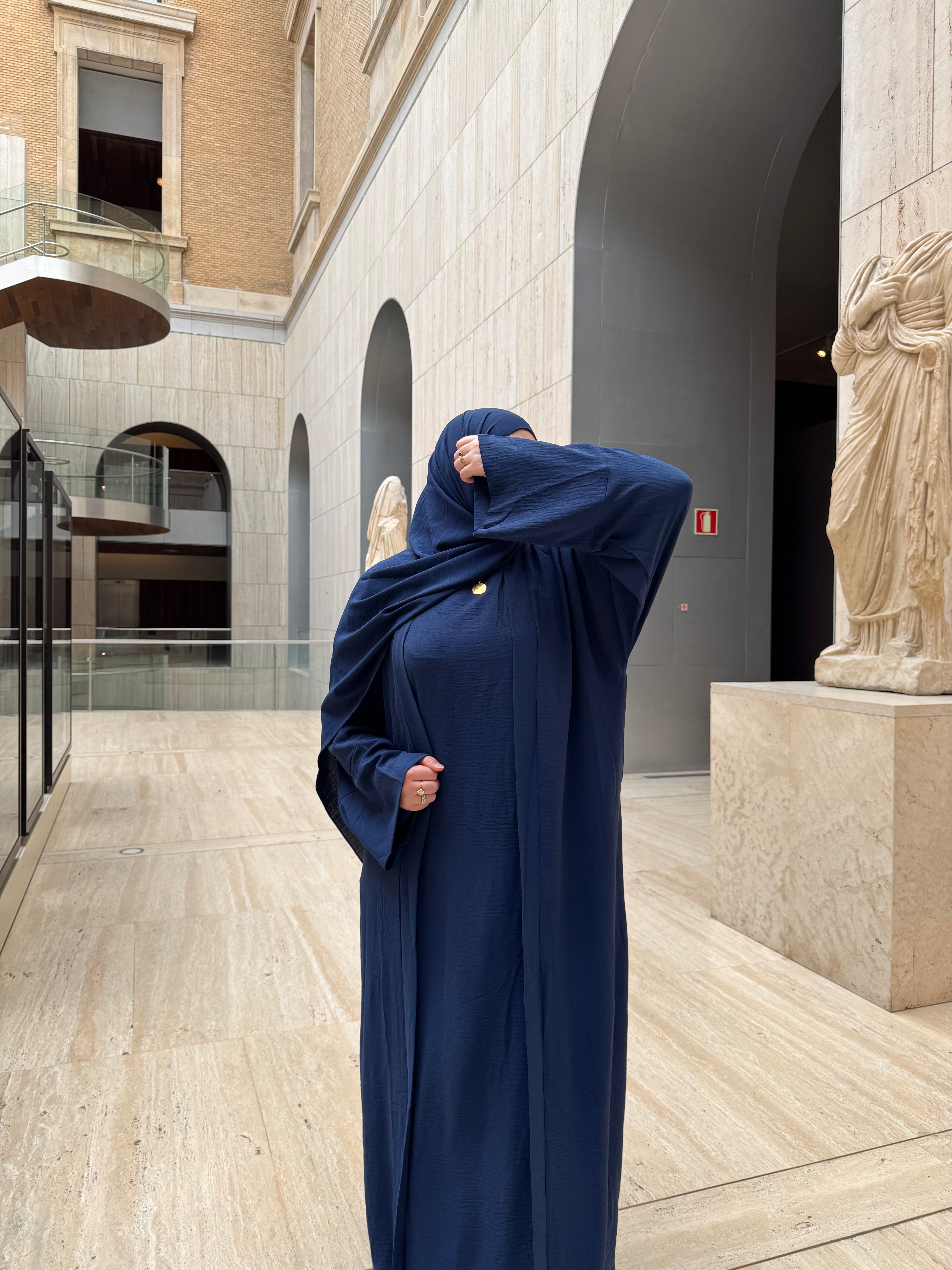 A person wearing a long navy abaya and matching hijab stands in a grand indoor courtyard with stone arches and classical statues, holding the hijab up near the face.