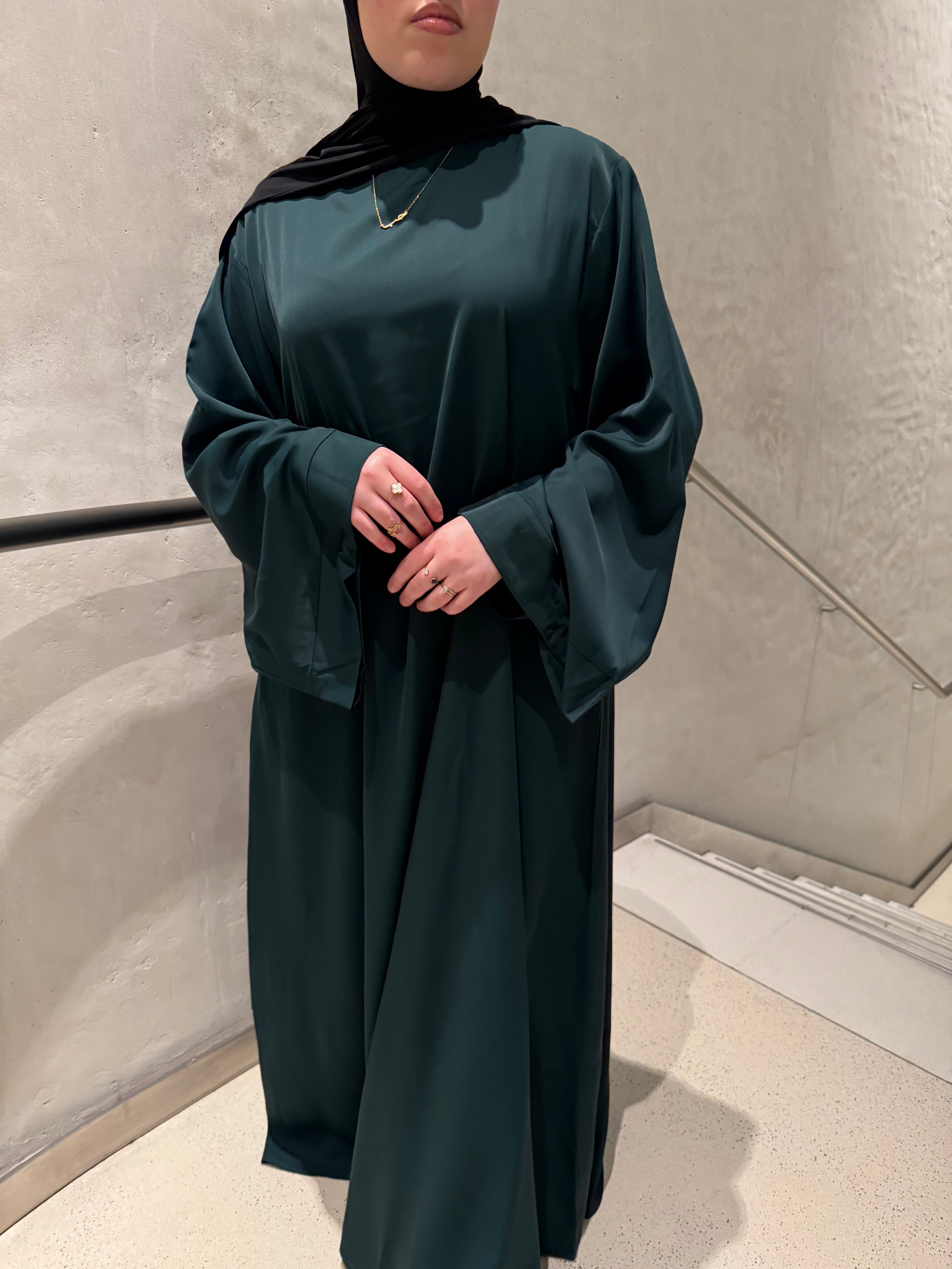A woman wearing a dark forest green abaya with wide flowing sleeves and a matching black hijab, standing indoors against a textured light gray wall.