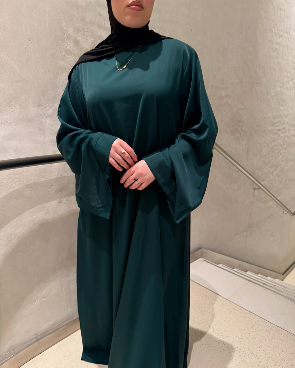A woman wearing a dark teal green abaya with wide bell sleeves and a matching black hijab, standing indoors against a textured light gray wall.