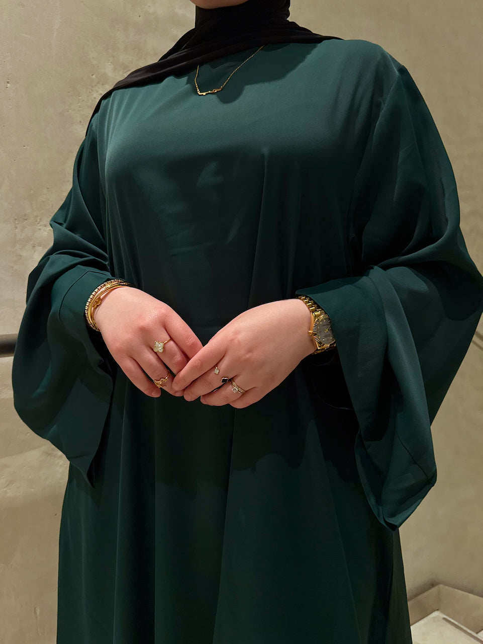 A person wearing a dark teal green abaya with wide draped sleeves, a black hijab, and gold jewelry, standing against a neutral textured wall.