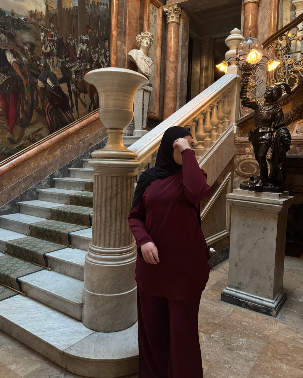 A woman wearing a deep burgundy long-sleeve maxi dress and black hijab stands in a grand, historic interior with marble stairs, classical statues, and a large mural.