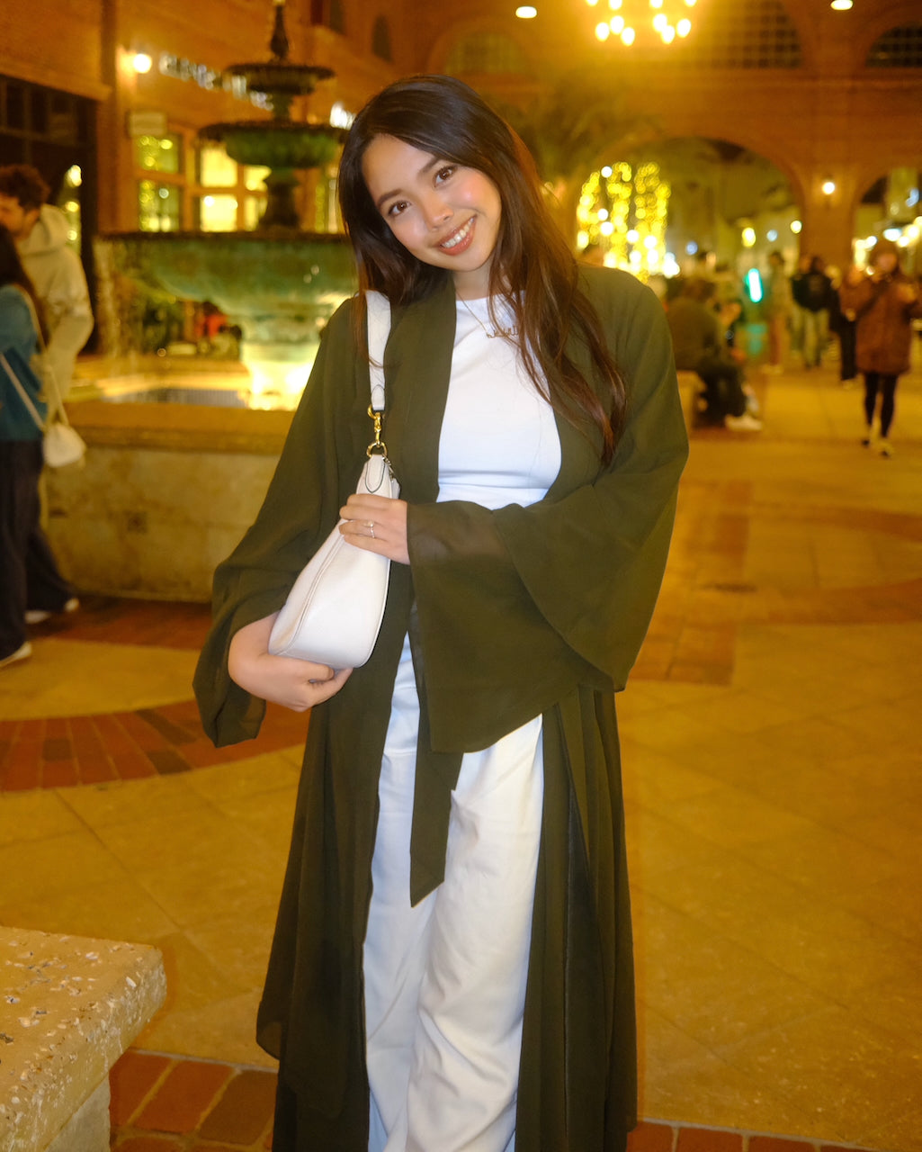 A woman posing in a warm, lit-up courtyard with a fountain in the background, wearing a dark olive green open-front cardigan over a white top and white pants, holding a white shoulder bag.