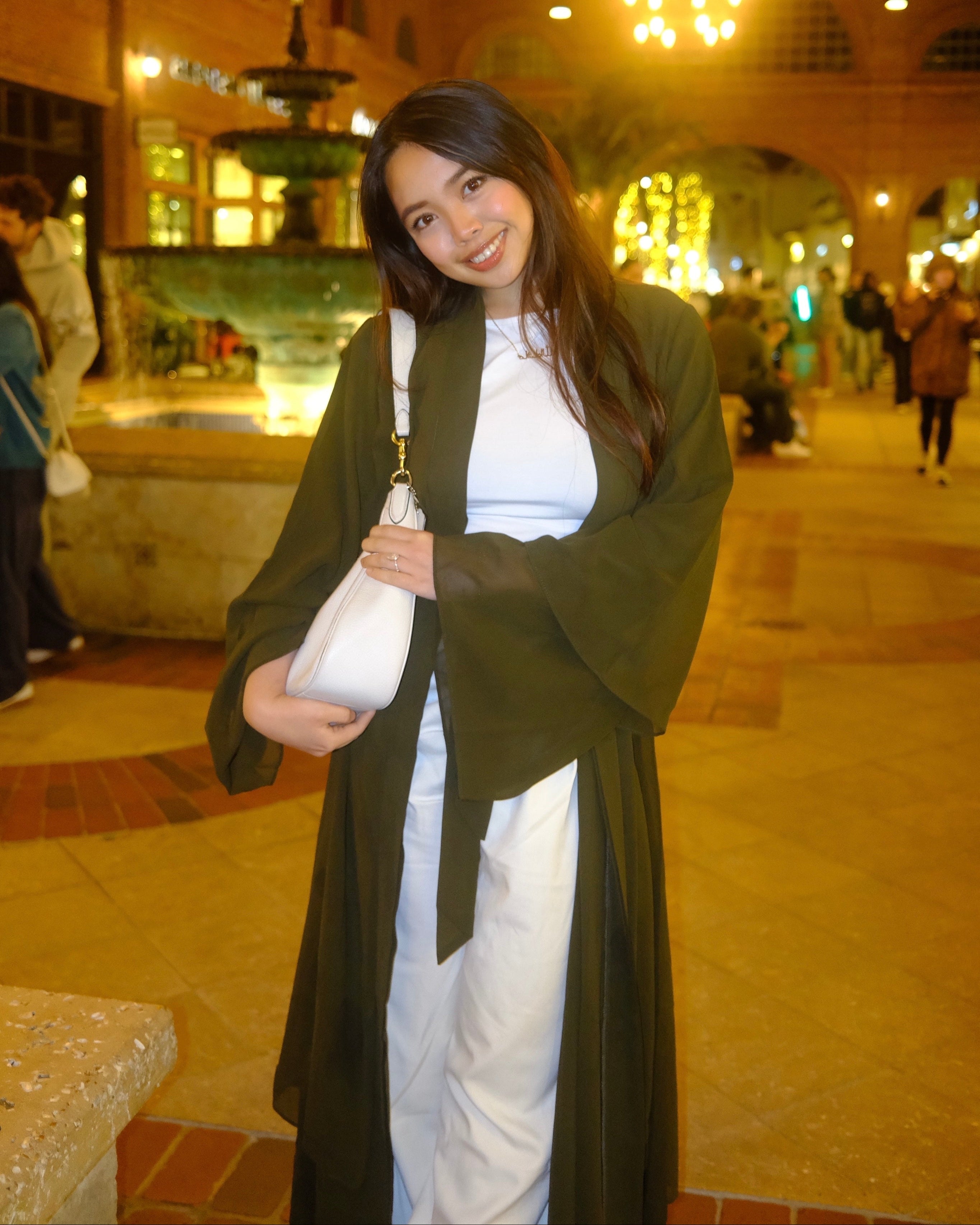 A woman posing in a warm, indoor architectural setting at night, wearing a white dress with a long dark olive green cardigan and holding a white shoulder bag.