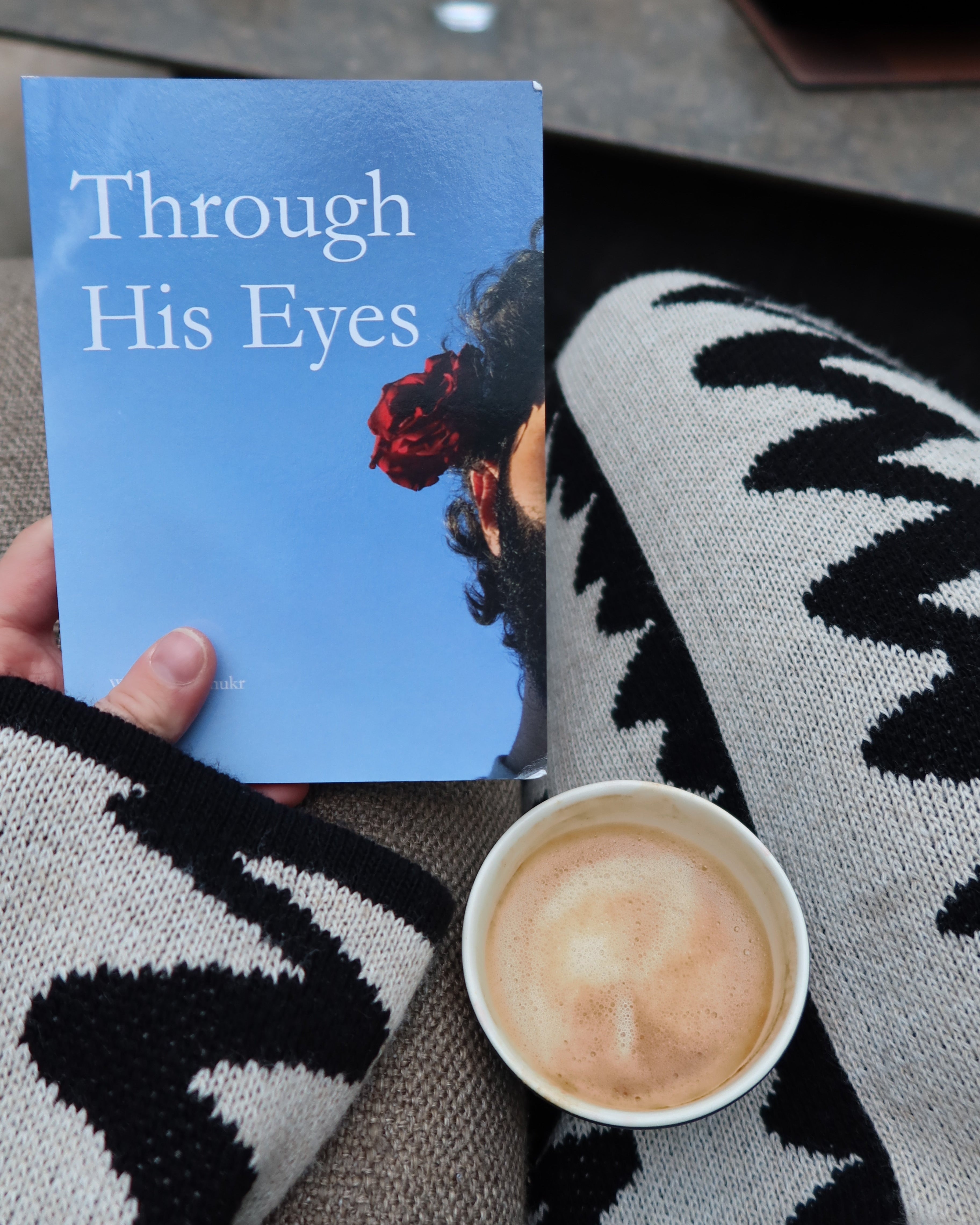 A hand holds a book titled 'Through His Eyes' with a blue cover, resting on a person's lap wrapped in a black and white patterned knit sweater, next to a cup of coffee.