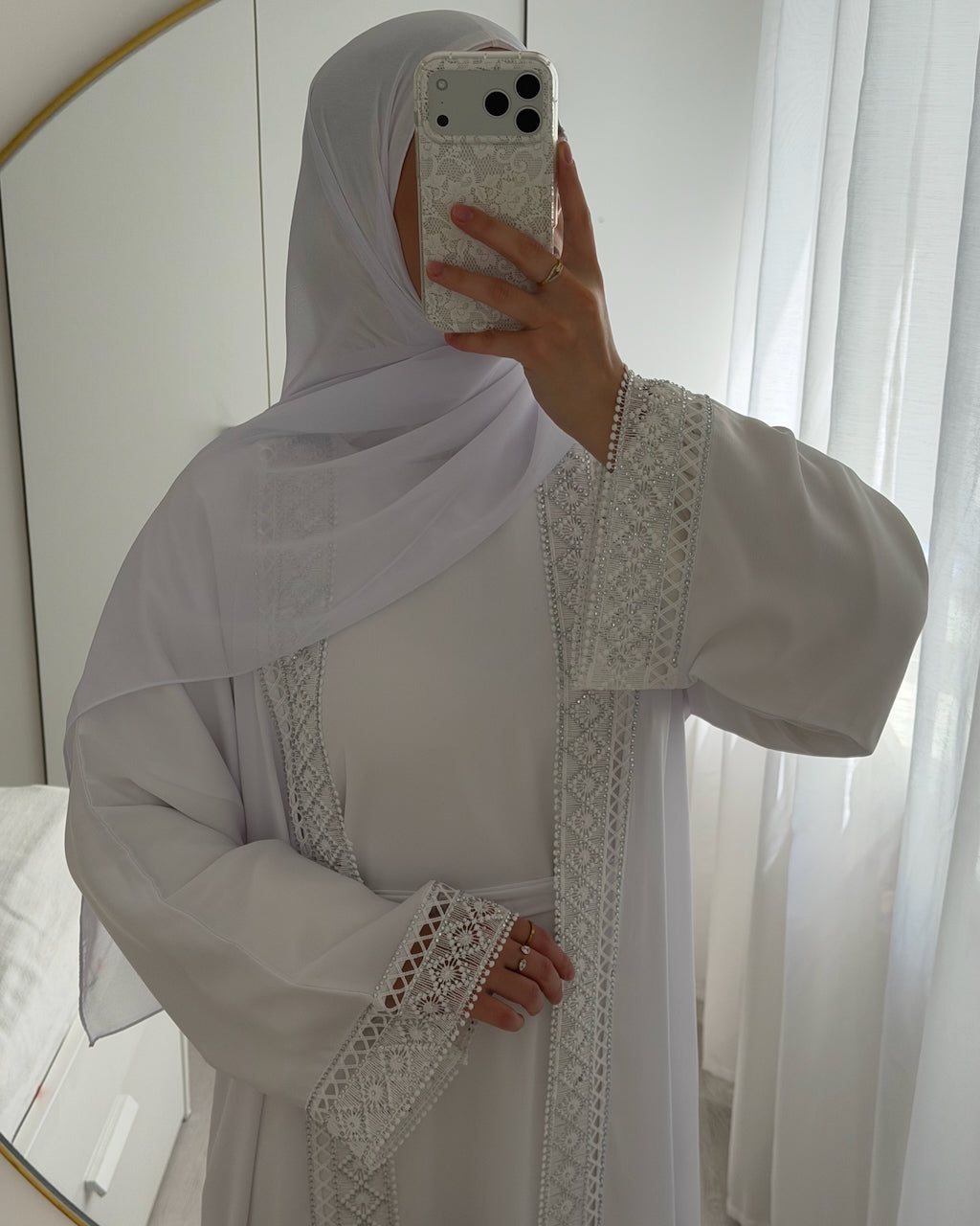 A woman wearing a white modest outfit with a matching hijab and a long white abaya featuring intricate silver embroidery, taking a mirror selfie in a bright room with white curtains. Modest Fashion - Fajr Noor