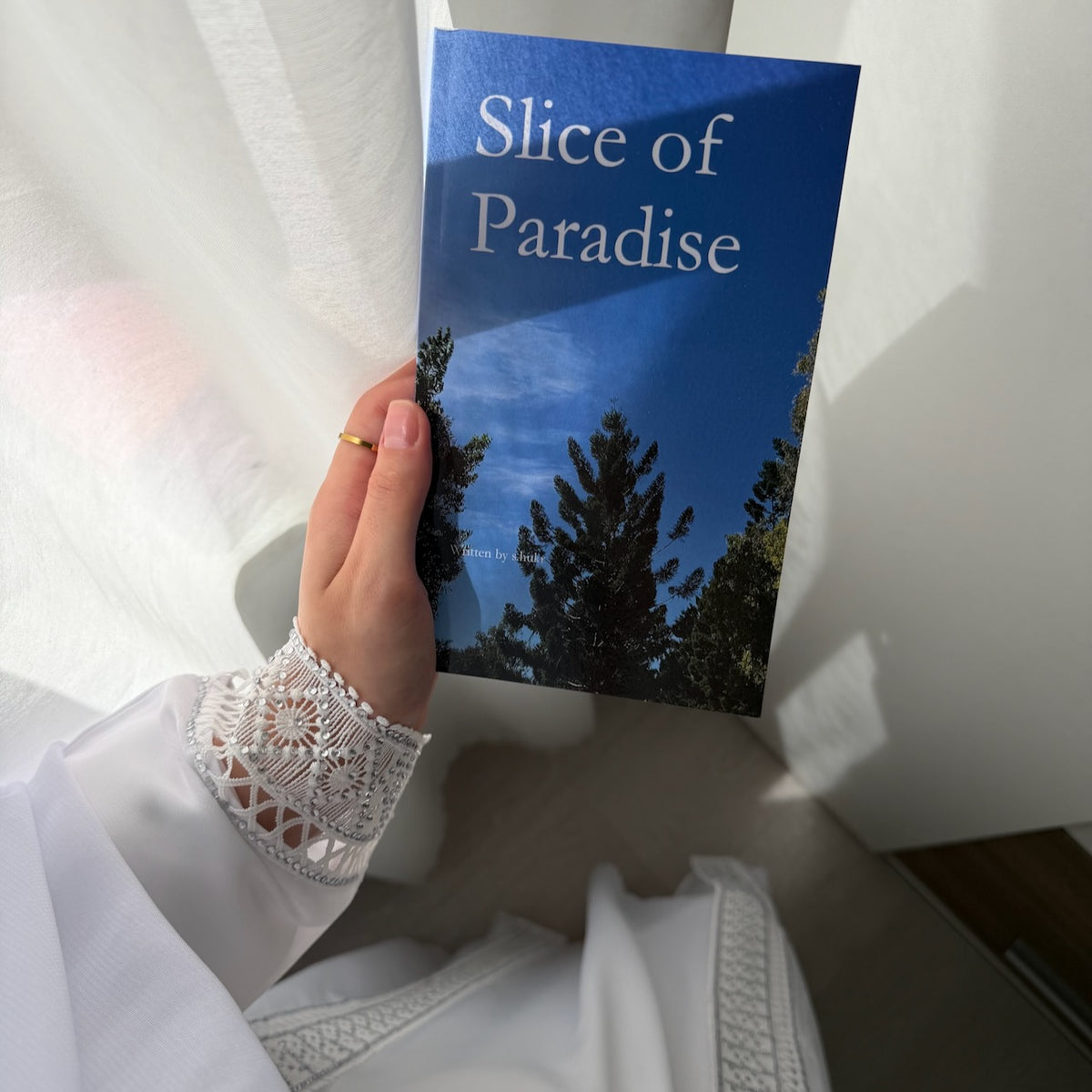 A hand holding a book titled 'Slice of Paradise' with a blue sky and tree cover illustration on the front, set against a soft white curtain background.